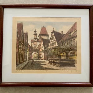 May include: Framed print of a European town square with buildings, a clock tower, and a fountain. The print has a brown wooden frame and a white mat. The buildings are in shades of brown, red, and beige. The print is signed.
