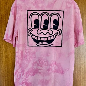 Puede incluir: Camiseta rosa te&ntilde;ida con un dise&ntilde;o de nudo con un gr&aacute;fico en blanco y negro de una cara sonriente con tres ojos. La camiseta tiene mangas cortas y cuello redondo. El dise&ntilde;o est&aacute; centrado en la parte posterior de la camiseta.
