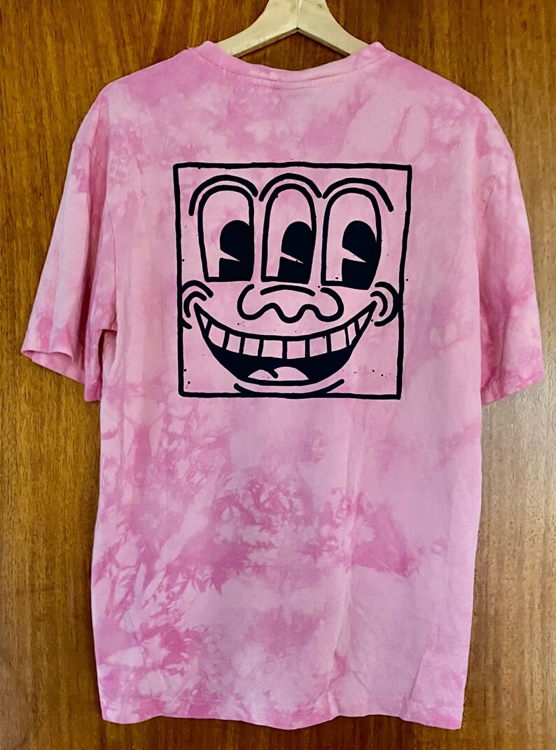 Puede incluir: Camiseta rosa te&ntilde;ida con un dise&ntilde;o de nudo con un gr&aacute;fico en blanco y negro de una cara sonriente con tres ojos. La camiseta tiene mangas cortas y cuello redondo. El dise&ntilde;o est&aacute; centrado en la parte posterior de la camiseta.