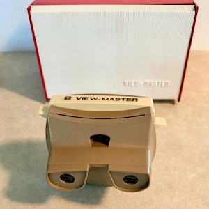 May include: A vintage View-Master viewer in tan and cream colors, with the "VIEW-MASTER" logo. It is paired with a red and white box. The viewer has two eye pieces and a central viewing area.