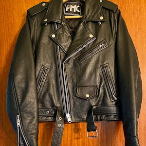 May include: Black leather motorcycle jacket with silver zippers, snaps, and a belt. The jacket has a classic asymmetrical design with a notched collar, shoulder epaulets, and a small pocket. The label reads "FMC".