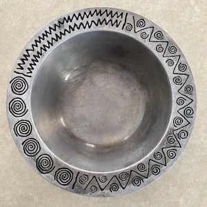 May include: A silver-colored metal bowl with a decorative black spiral and zigzag pattern around the rim. The bowl has a simple, rounded shape and a smooth interior.