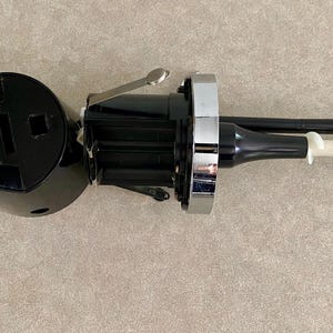 May include: Black and silver wine opener with a white corkscrew. The opener has a circular base with text and a square opening. The handle is black with a silver accent. The corkscrew is white.