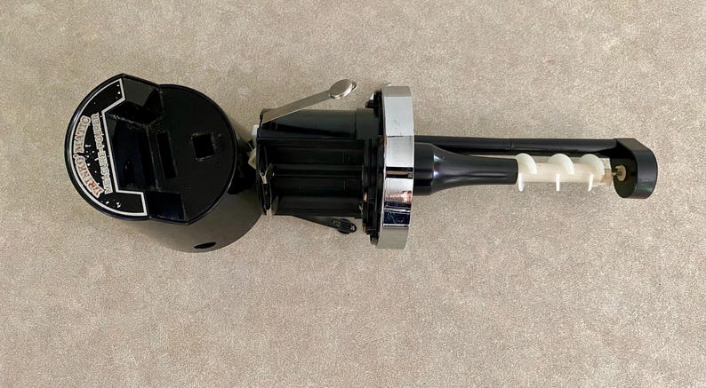 May include: Black and silver wine opener with a white corkscrew. The opener has a circular base with text and a square opening. The handle is black with a silver accent. The corkscrew is white.