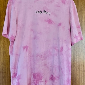 Puede incluir: Camiseta rosa te&ntilde;ida con la firma "Keith Haring" en escritura negra. La camiseta est&aacute; colgada en una percha de madera. La camiseta tiene cuello redondo y mangas cortas.
