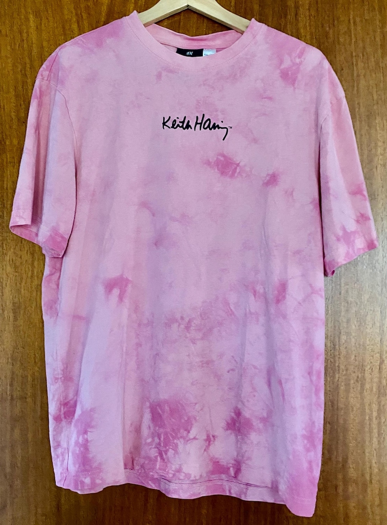 Puede incluir: Camiseta rosa te&ntilde;ida con la firma "Keith Haring" en escritura negra. La camiseta est&aacute; colgada en una percha de madera. La camiseta tiene cuello redondo y mangas cortas.