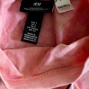 Puede incluir: Primer plano de una camiseta rosa con una etiqueta negra de H&M. La etiqueta incluye el texto "RELAXED FIT COUPE D&Eacute;CONTRACTEE" e informaci&oacute;n sobre la talla. Tambi&eacute;n es visible una etiqueta blanca m&aacute;s peque&ntilde;a con el nombre "Keta Han".