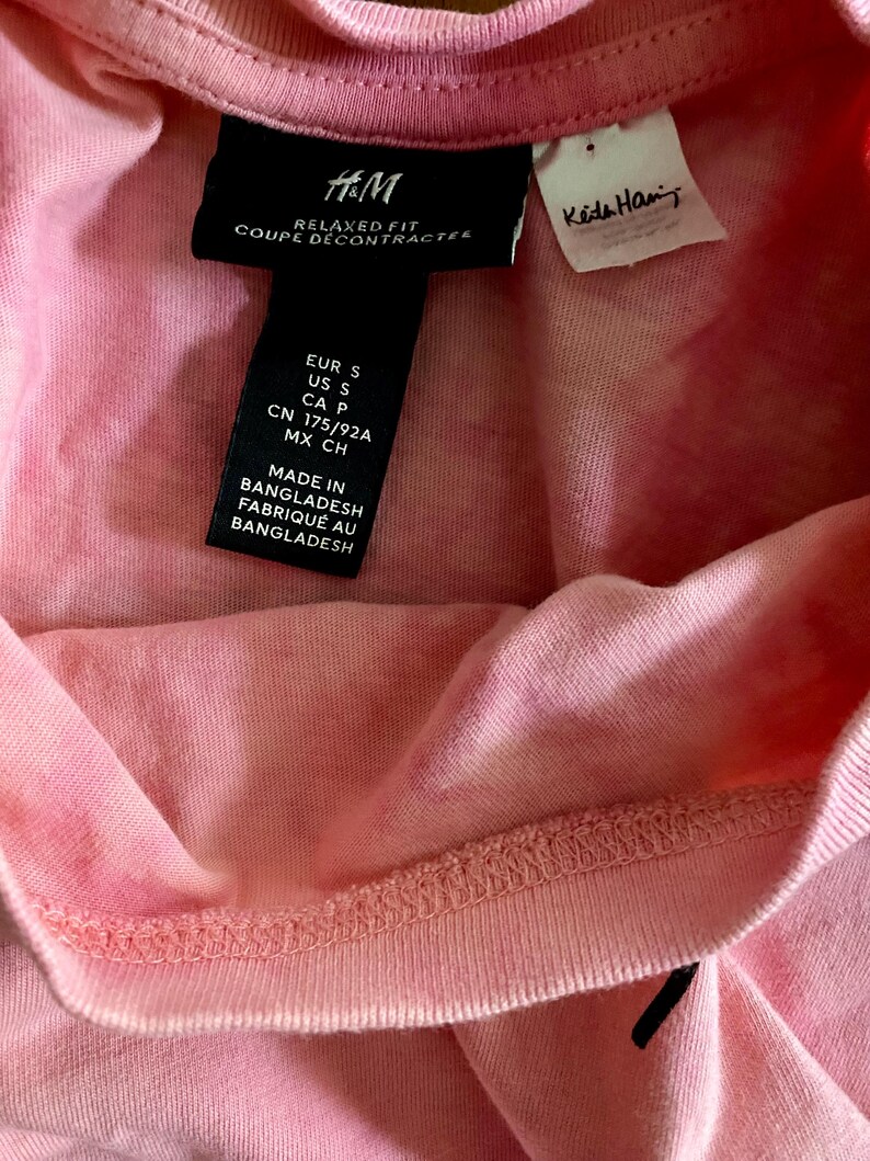 Puede incluir: Primer plano de una camiseta rosa con una etiqueta negra de H&M. La etiqueta incluye el texto "RELAXED FIT COUPE D&Eacute;CONTRACTEE" e informaci&oacute;n sobre la talla. Tambi&eacute;n es visible una etiqueta blanca m&aacute;s peque&ntilde;a con el nombre "Keta Han".