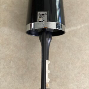 May include: A black and silver bar tool with the word "FORDA" on the front. The tool has a cylindrical top, a silver band, and a long, black stem with a white plastic piece. The base is a black, hexagonal shape.