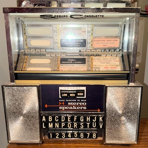 May include: Vintage Seeburg Consoltte jukebox with chrome accents. Features selection buttons, volume control, and song title displays. The jukebox has a glass front and a dark blue control panel. The text on the jukebox includes "stereo speakers" and "Hot Stuff".