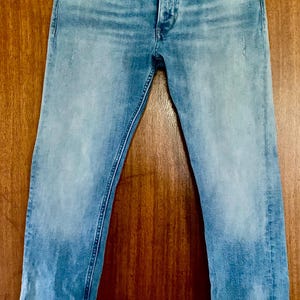 May include: Light blue denim jeans with a button fly and five-pocket design. The jeans have a faded wash and the brand name "Calvin Klein Jeans" is visible on the waistband. The jeans are hanging on a wooden hanger.