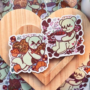 May include: Two adorable green bear stickers with glitter accents. One bear is holding a cup of coffee and the other is holding a croissant. Both stickers have a brown and white background with coffee beans and other details.