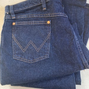 May include: Dark blue denim jeans folded neatly. The jeans feature a classic five-pocket design with a back pocket showcasing a distinctive 'W' stitch in a contrasting tan thread. Copper rivets and tan stitching detail the seams.