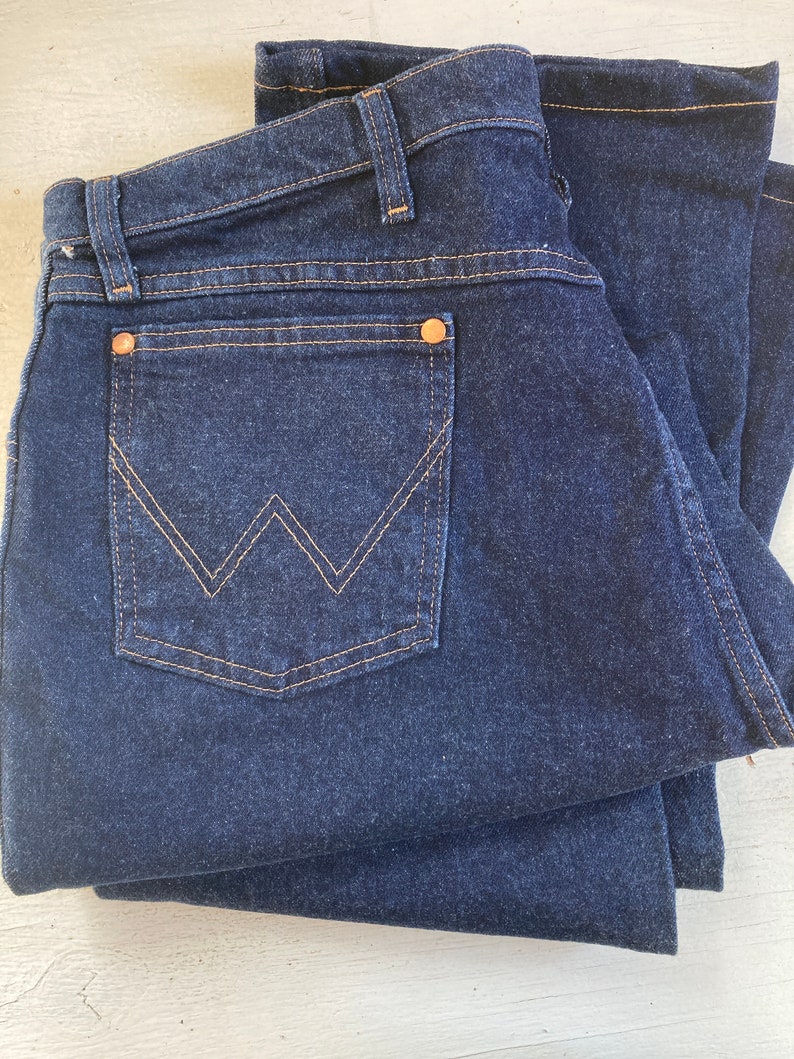 May include: Dark blue denim jeans folded neatly. The jeans feature a classic five-pocket design with a back pocket showcasing a distinctive 'W' stitch in a contrasting tan thread. Copper rivets and tan stitching detail the seams.