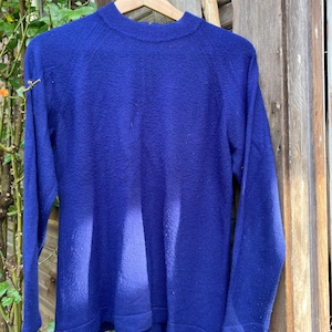 May include: A royal blue sweater with long sleeves and a mock neck. The sweater is hanging on a wooden hanger against a weathered wooden background. The sweater's texture appears soft and slightly fuzzy.
