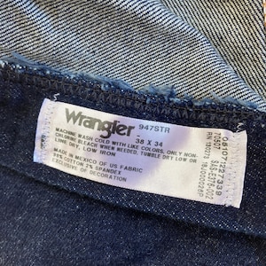 May include: Close-up of a dark blue denim fabric with a white label. The label reads "Wrangler 947STR" and indicates the size as 38 x 34 inches. The fabric is made in Mexico of US fabric, 98% cotton, and 2% spandex.