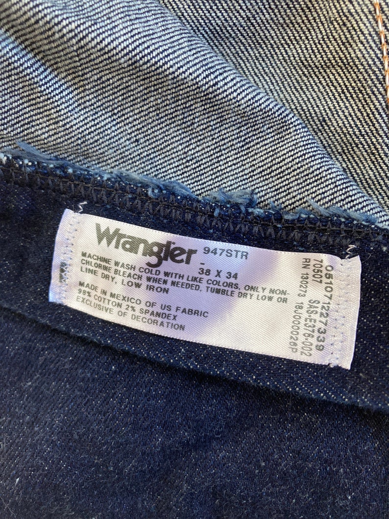 May include: Close-up of a dark blue denim fabric with a white label. The label reads "Wrangler 947STR" and indicates the size as 38 x 34 inches. The fabric is made in Mexico of US fabric, 98% cotton, and 2% spandex.