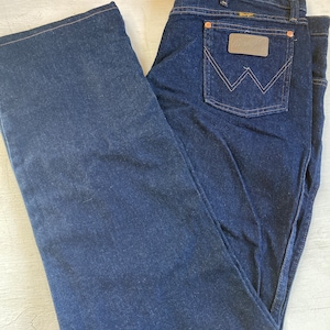 May include: Dark blue denim jeans with a classic five-pocket design. Features a brown leather patch on the back pocket with the Wrangler logo. The jeans have a straight leg cut and are folded neatly.