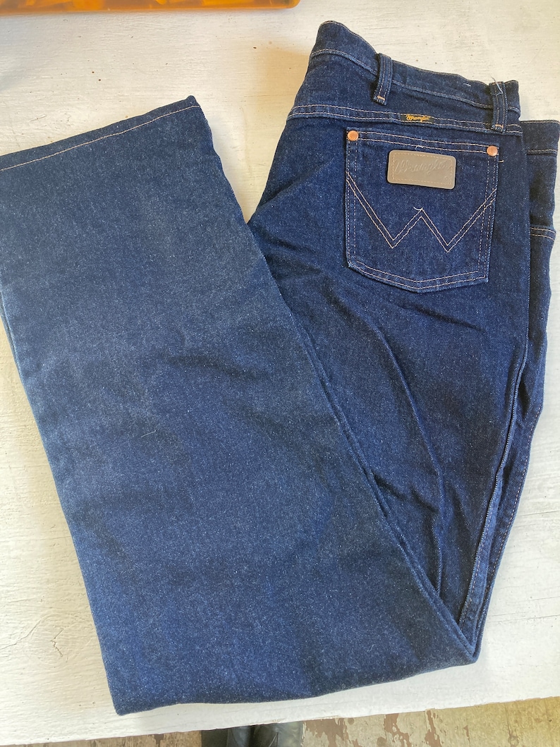 May include: Dark blue denim jeans with a classic five-pocket design. Features a brown leather patch on the back pocket with the Wrangler logo. The jeans have a straight leg cut and are folded neatly.