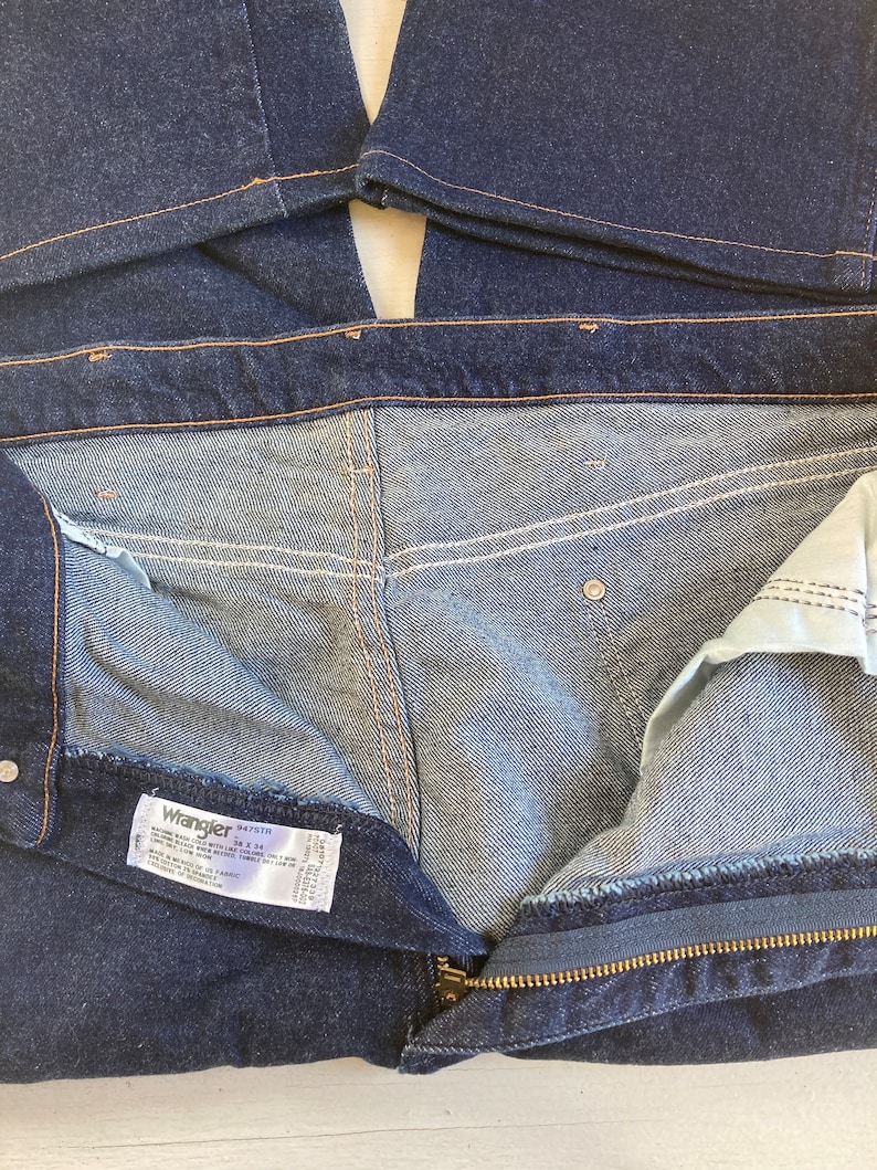 May include: Dark blue denim jeans with a zipper fly and a visible label. The jeans have a classic five-pocket design and are made of a durable denim fabric. The stitching is a contrasting orange color.