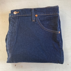 May include: Folded dark blue denim jeans with copper-colored rivets and a button. The jeans have a classic five-pocket design with tan stitching. The brand name is visible on the button.