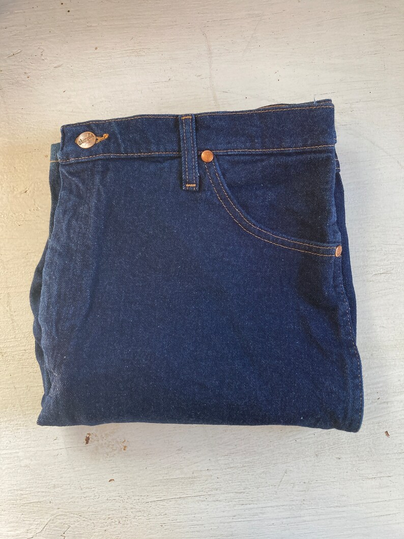 May include: Folded dark blue denim jeans with copper-colored rivets and a button. The jeans have a classic five-pocket design with tan stitching. The brand name is visible on the button.