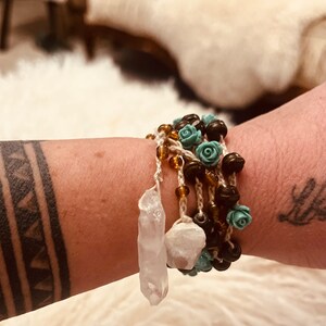 Op de afbeelding: Een gelaagde armband met bruine houten kralen, turquoise bloemenaccenten en een witte kristallen hanger.