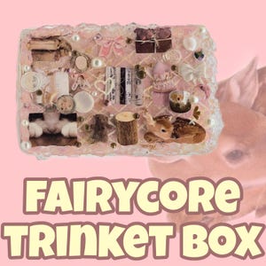 May include: A rectangular trinket box with a pink and white design, adorned with miniature decorative elements. The box features a variety of small items, including a miniature deer, bows, pearls, and tiny books. The text "FAIRYCORE TRINKET BOX" is displayed at the bottom.