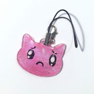 May include: A pink, glittery cat-shaped charm with a sad expression. The charm has black eyes and a curved mouth, attached to a black cord with a silver clasp. The background is plain white.