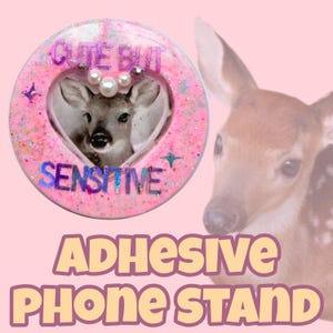 May include: A pink adhesive phone stand with a deer image inside a heart-shaped frame. The frame has the words "CUTE BUT" and "SENSITIVE" in holographic lettering. The stand is decorated with glitter and faux pearls.