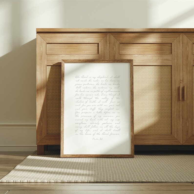 May include: A rectangular wall art print in a brown wooden frame featuring the text of Psalm 23 written in a light gray cursive script on a white background. The print is positioned upright, displaying the full biblical passage clearly.