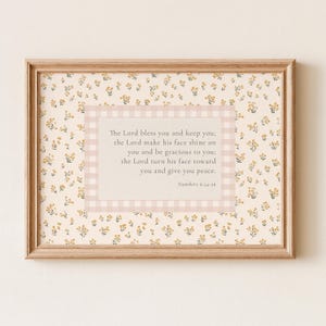 Op de afbeelding: Een ingelijste wandkunstprint met een lichtcrème achtergrond en een herhalend patroon van kleine gele bloemen. Een centraal rechthoekig blok met een lichtroze gingham-rand bevat de tekst: The Lord bless you and keep you; the Lord make his face shine on you and be gracious to you; the Lord turn his face toward you and give you peace. Numbers 6:24-26.