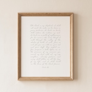 May include: A framed wall art print featuring the text of Psalm 23 written in a delicate, light gray cursive script on a white background. The frame is made of light-toned wood with a simple, clean profile, surrounding the white mat and print.