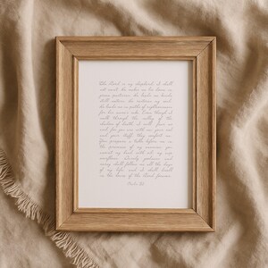 May include: A rectangular wooden frame with a light brown grain finish containing a white print featuring the text of Psalm 23 in a grey cursive script. The frame has a simple, clean design with mitered corners and a flat, smooth surface profile.