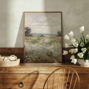 May include: A framed landscape print with a field of white flowers and trees. The print has the text "Give thanks to the Lord, for He is good; His love endures forever. 1. Corinthians 16:14". The print is displayed on a wooden dresser with a vase of white tulips.