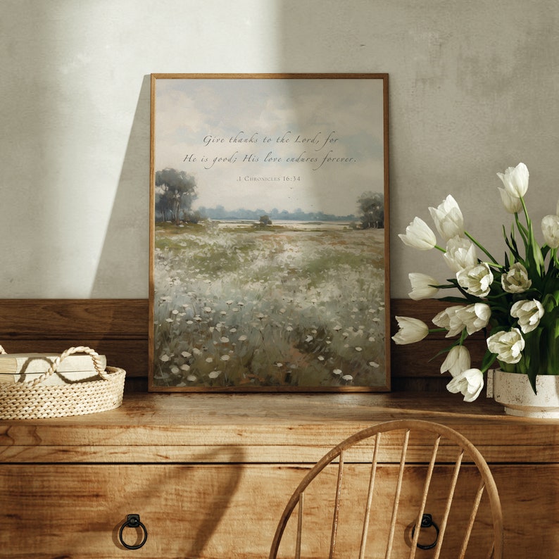 May include: A framed landscape print with a field of white flowers and trees. The print has the text "Give thanks to the Lord, for He is good; His love endures forever. 1. Corinthians 16:14". The print is displayed on a wooden dresser with a vase of white tulips.