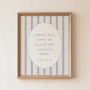 May include: A framed wall art print featuring a beige background with vertical blue stripes. Centered is an oval containing the text: I PRAISE YOU, FOR I AM fearfully AND wonderfully MADE. PSALM 139:14. The frame is made of light brown wood.
