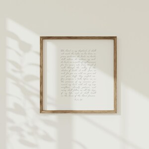 May include: A square wall art piece featuring a wooden frame and a white background with Psalm 23 written in dark grey cursive script. The text reads: The Lord is my shepherd; I shall not want. He makes me lie down in green pastures.