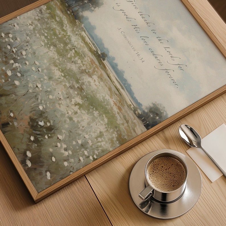 May include: A framed landscape art print with a field of white flowers and the text "Give thanks to the Lord, for is good; His love endures forever. 1 Chronicles 16:34." A silver coffee cup and saucer with coffee and a spoon are on a wooden table.