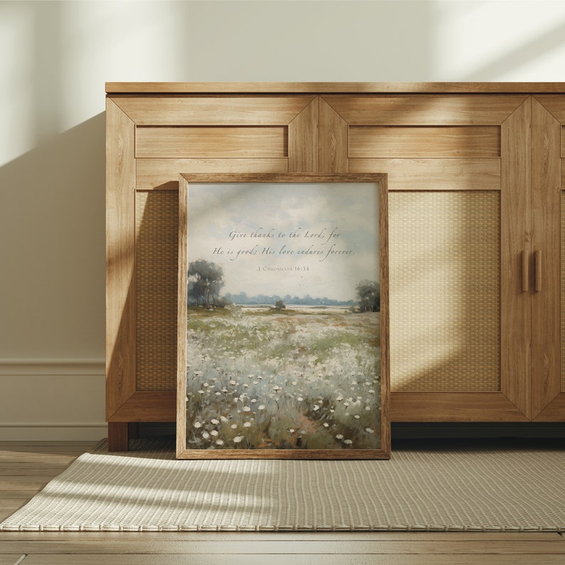 May include: A framed landscape art print with a wooden frame. The artwork depicts a field of white flowers under a cloudy sky. The text "Give thanks to the Lord, for He is good; His love endures forever. 1 Chronicles 16:34" is printed at the top.