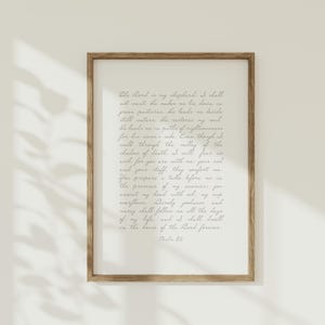 May include: A framed wall art print featuring Psalm 23 written in a dark gray cursive script on a light cream background. The text begins with The Lord is my shepherd and concludes with Psalm 23. The frame is made of medium-toned brown wood.