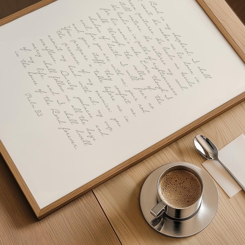 May include: A framed print featuring the text of Psalm 23 in black cursive script on a white background. Beside the frame sits a stainless steel cup filled with coffee on a matching saucer, accompanied by a small silver spoon on a white napkin.