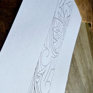 Op de afbeelding: Een zwart-witte lijntekening van een bloemmotief met een zonnebloem in het midden. Het ontwerp is op een witte achtergrond.