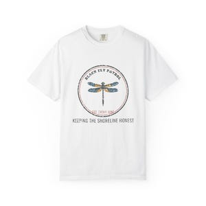 Może przedstawiać: Biały t-shirt z krótkim rękawem z okrągłą grafiką przedstawiającą ważkę. Napis głosi BLACK FLY PATROL, EST. EVERY JUNE oraz KEEPING THE SHORELINE HONEST. Wzór ma postarzany, przetarty wygląd.