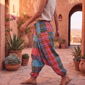 May include: A person wearing a beige tank top and colorful plaid harem pants with a drawstring waist. The pants feature a mix of red, orange, blue, and green stripes. Brown sandals complete the outfit. The setting is an outdoor walkway with terracotta pots and plants.