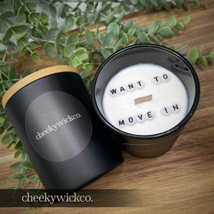 May include: Two black candle jars with wooden lids. One jar has a white label with the text "cheekywickco." The other jar has a white wax surface with black letter tiles spelling "WANT TO MOVE IN."