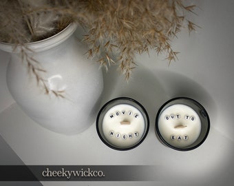 May include: Two black candle jars with white wax and wooden wicks. The wax has white letters spelling out "Movie Night" and "Out to Eat".