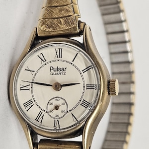Op de afbeelding: Een goudkleurige Pulsar quartz-horloge met een witte wijzerplaat en Romeinse cijfers voor de uren. Het horloge heeft een goudkleurige metalen band.