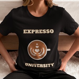 Puede incluir: Camiseta negra con el texto "Espresso University" en letras doradas. Una taza de café estilizada con corazones y una corona de laurel está centrada en la camiseta.