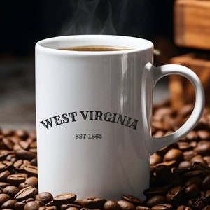 Puede incluir: Taza de café de cerámica blanca con el texto "West Virginia Est 1863" impreso en el lateral. La taza está llena de café y sale vapor de la parte superior. La taza está sobre un lecho de granos de café.